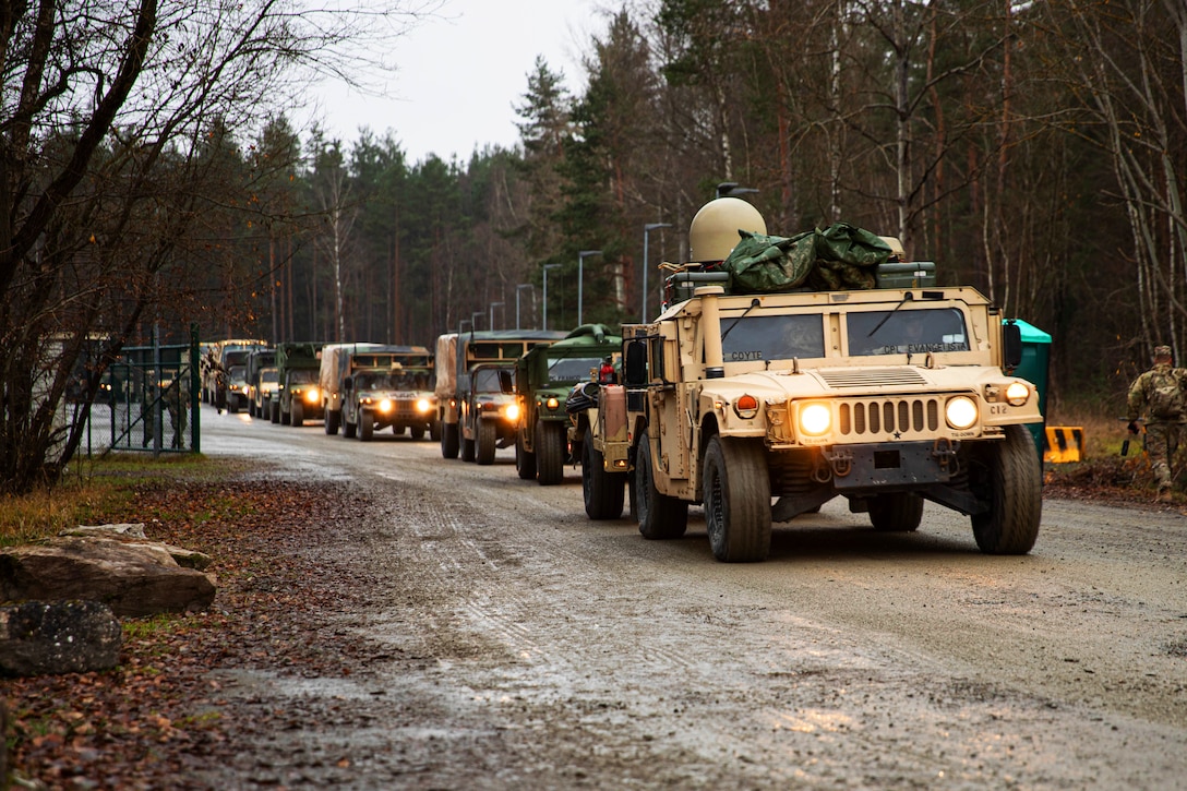 Army military vehicles drive in a line on a tree-lined roadway.