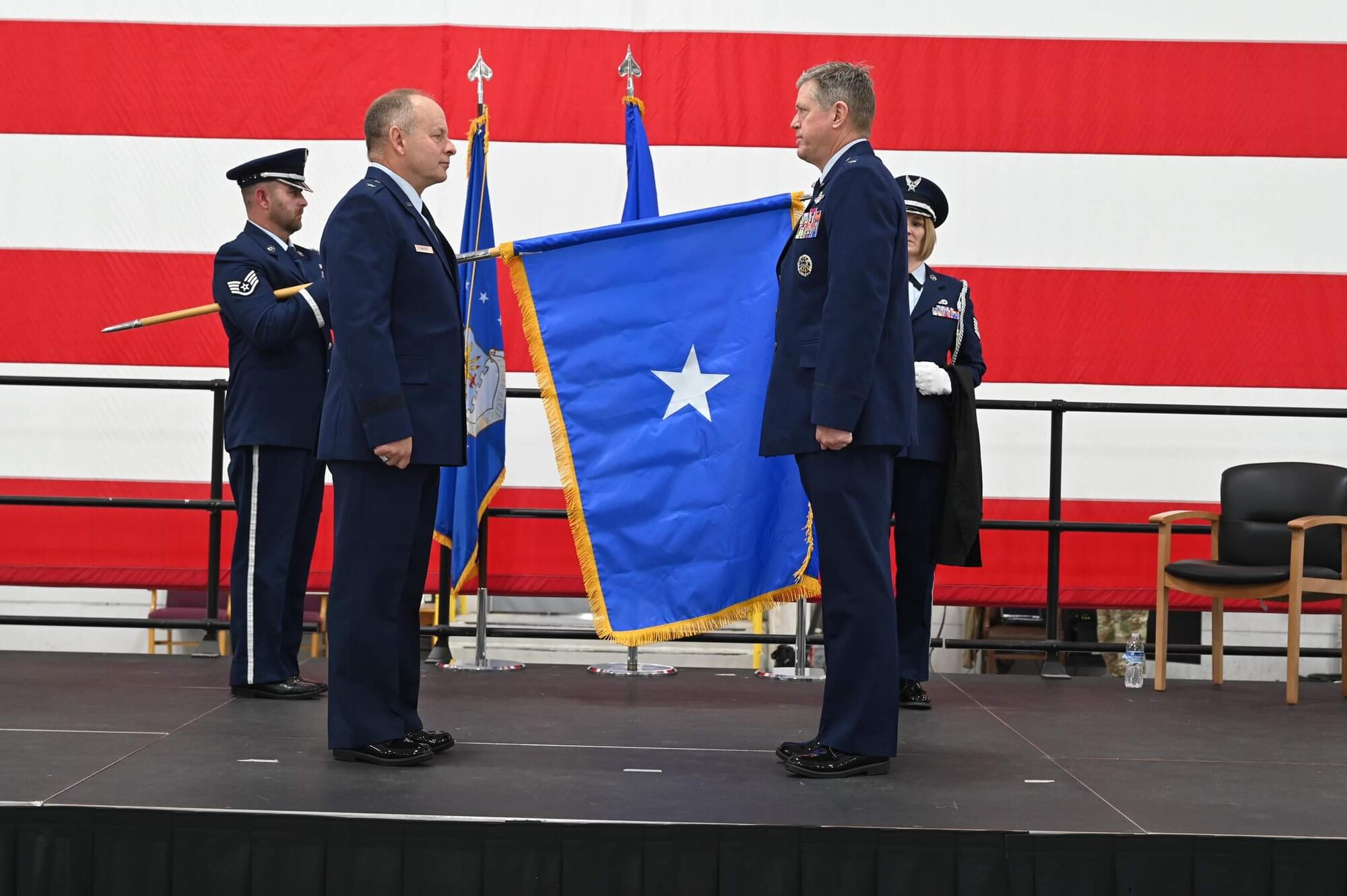 Two men in U.S. Air Force service dress face each other at attention in the foreground while a man in Air Force Honor Guard uniform holds a blue flag with a single white star unfurled in the background.