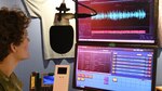An image of a radio broadcaster preparing material for a radio show. A studio microphone and computers with broadcasting and editing software are visible in the background.