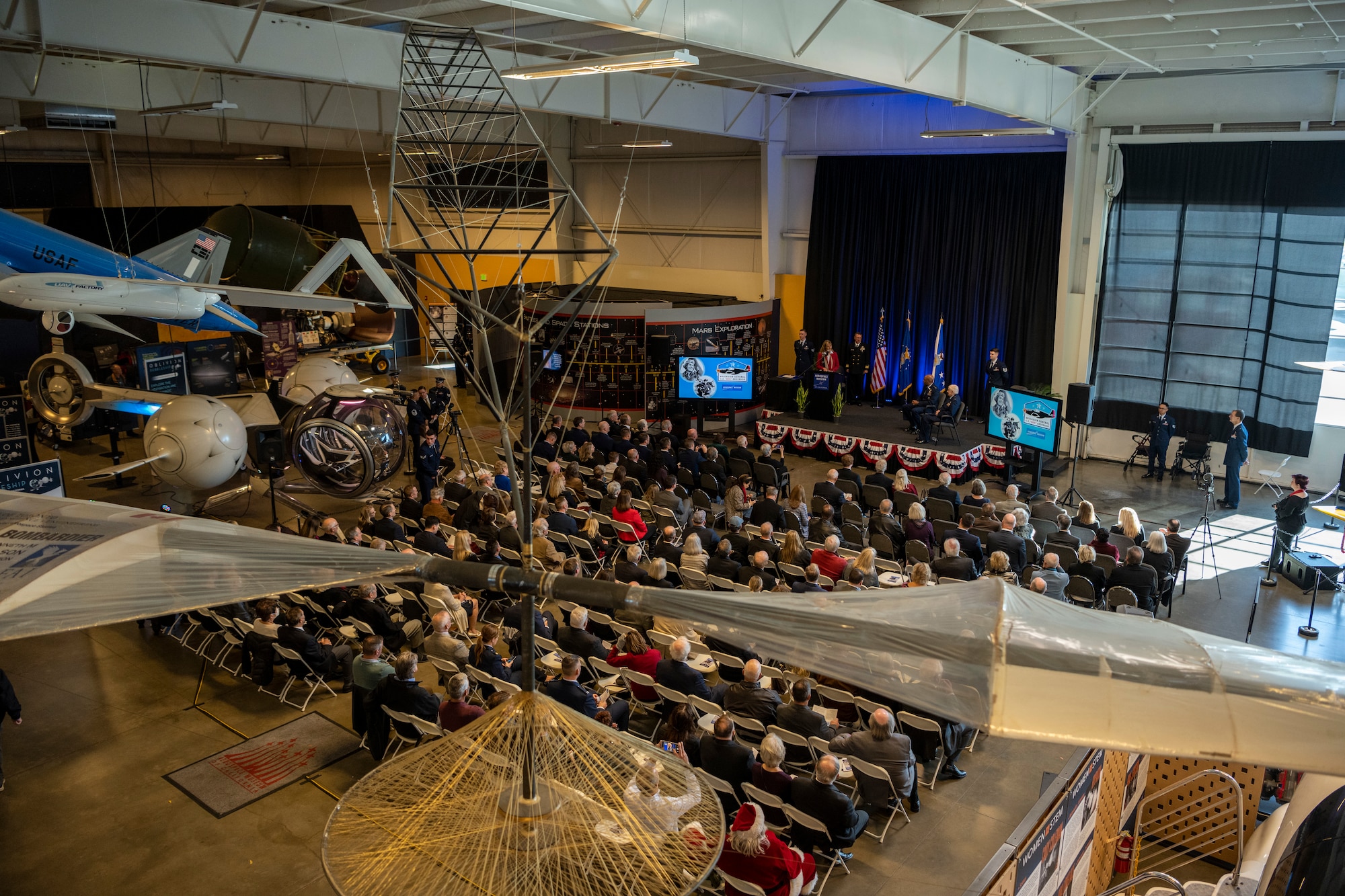 An overall photo of a large aviation museum with aircraft displayed throughout as well as a large crowd of people sitting in chairs in front of a stage.