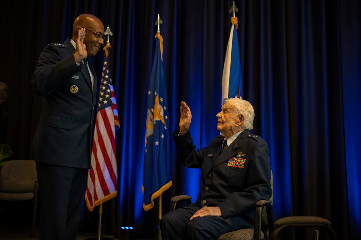 And elderly man sits in a chair and a high-ranking military leader stand, and they both have their arms raised like they are giving an oath.
