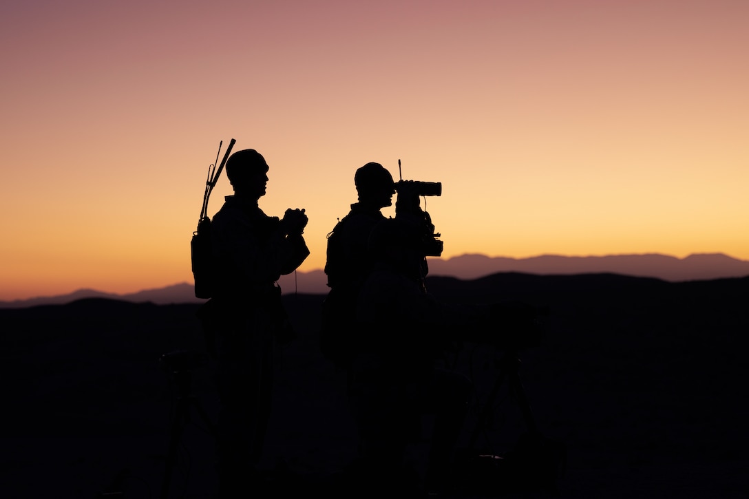 Two Marines use binoculars while standing in the dark as the sun peaks through.