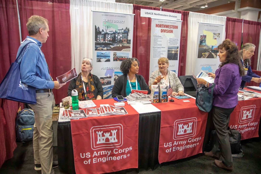 Photo of a group of people around an exhibit table draped with a tablecloth that reads "Army Corps of Engineers."