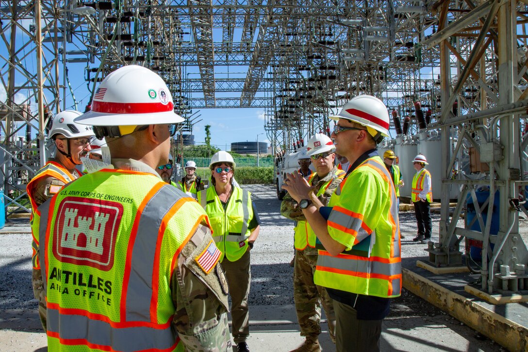 A group of US Army Corps of Engineers personnel in reflective vests and hard hats stand in a circle surrounded by electrical transformers and transmission equipment.