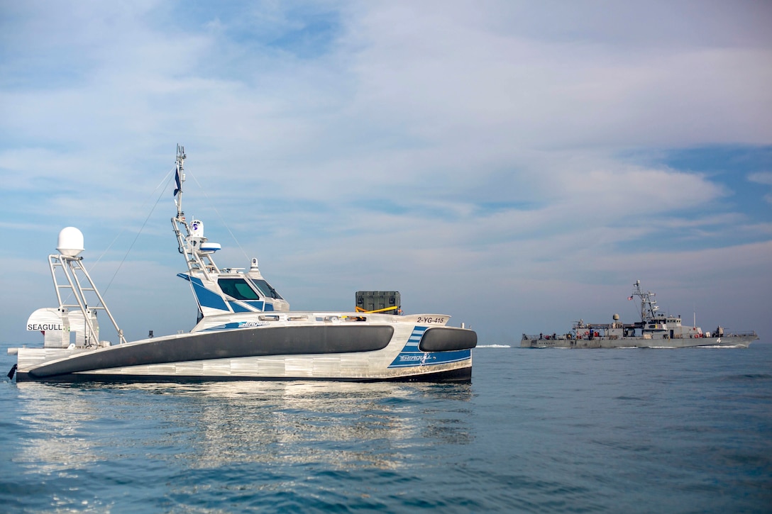 A small unmanned vessel sails in open water, with a larger ship in the background.