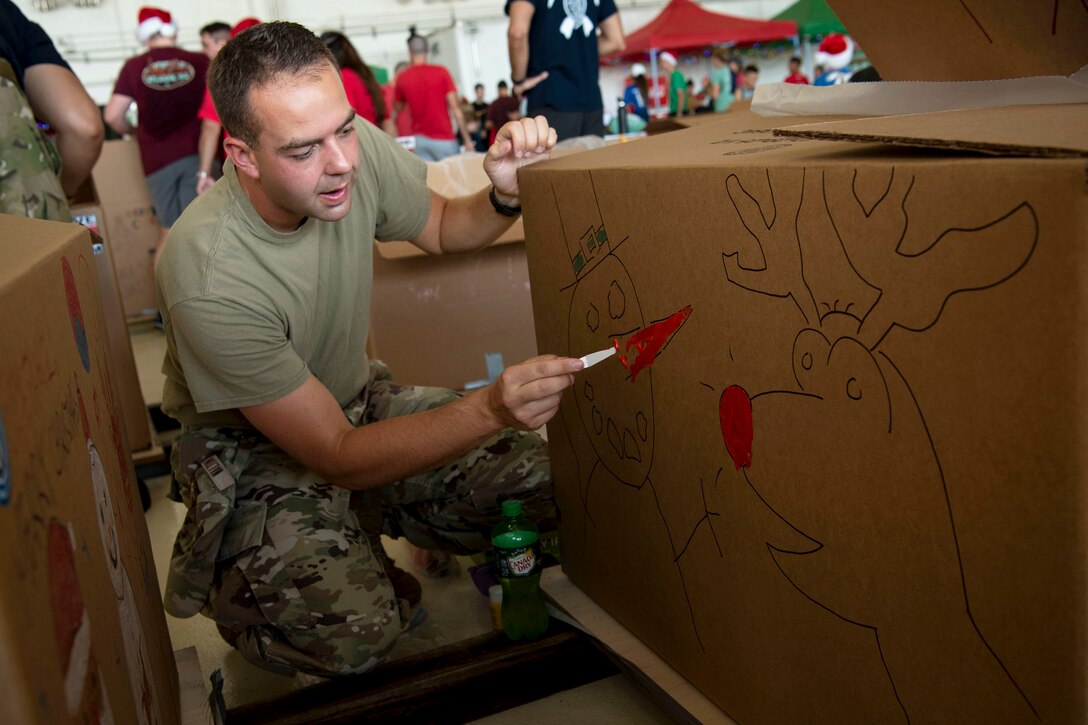 A kneeling airman colors the nose of a snowman drawn on a large cardboard box.