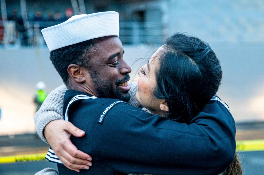 A smiling sailor and civilian embrace on a pier, a ship in the background.