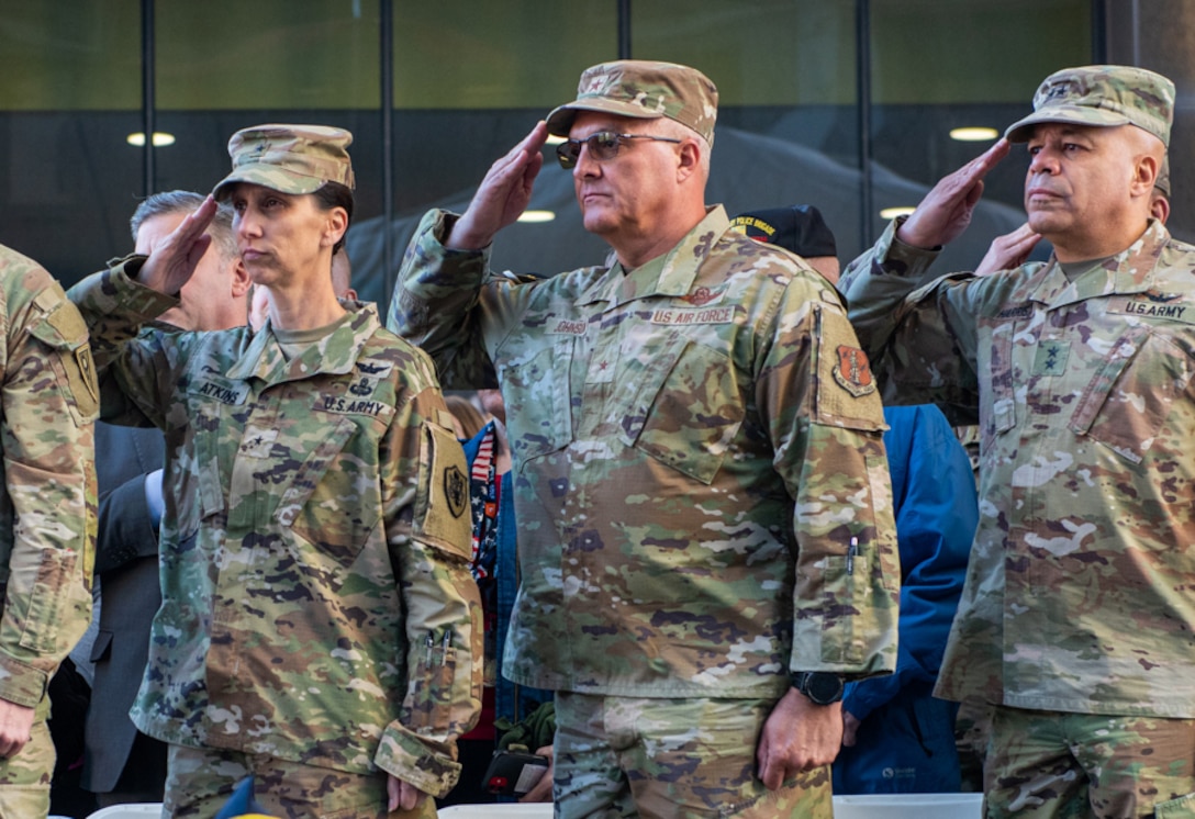 Three military members in camoflaged OCP uniforms salute on the viewing stand. Two men and one woman.
