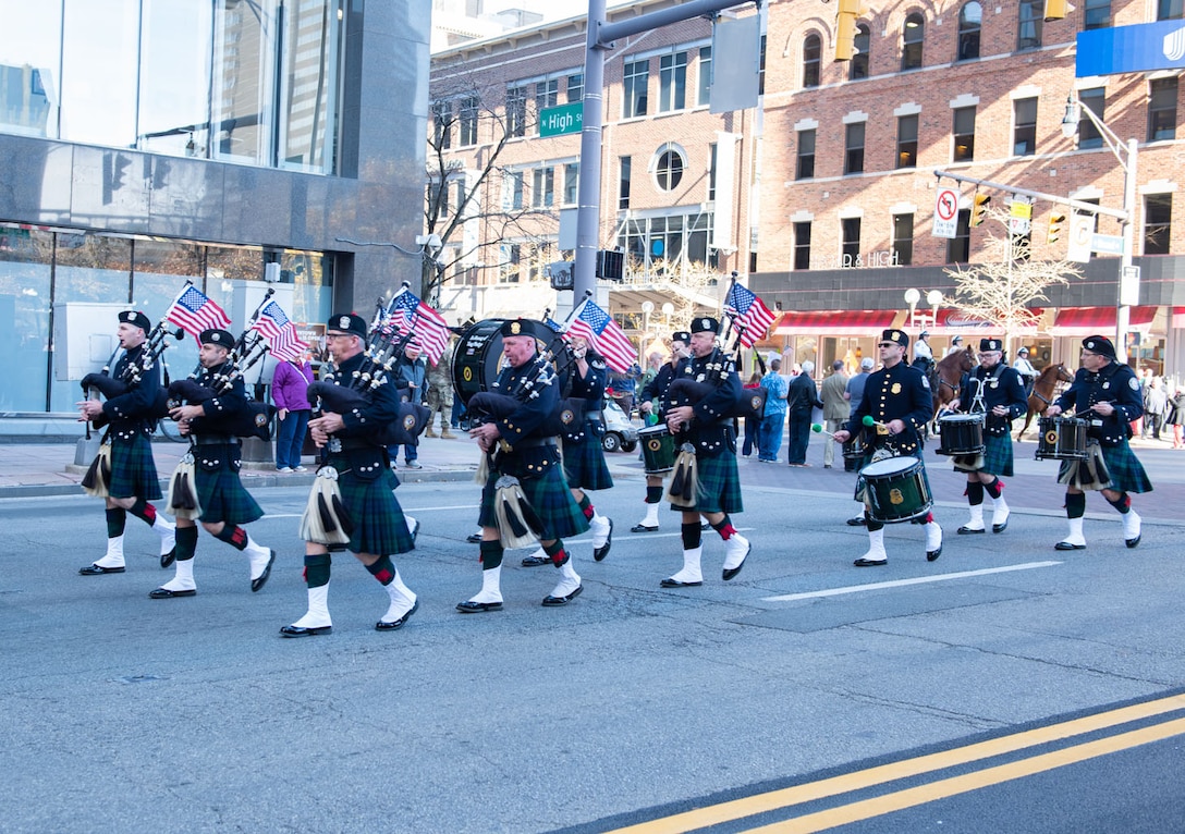 A police marching band in Scottish-type uniforms march down the street. Some have bagpipes, some have flutes and some have drums.
