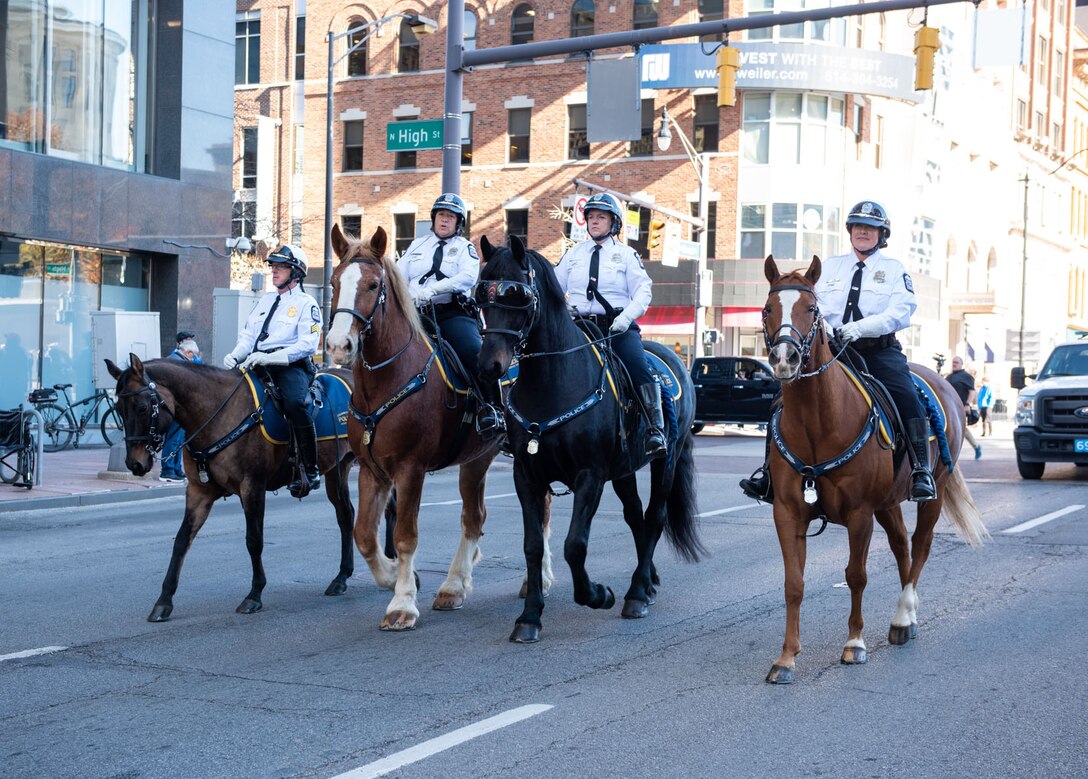 Four people in white and black police uniforms ride horses down the street.