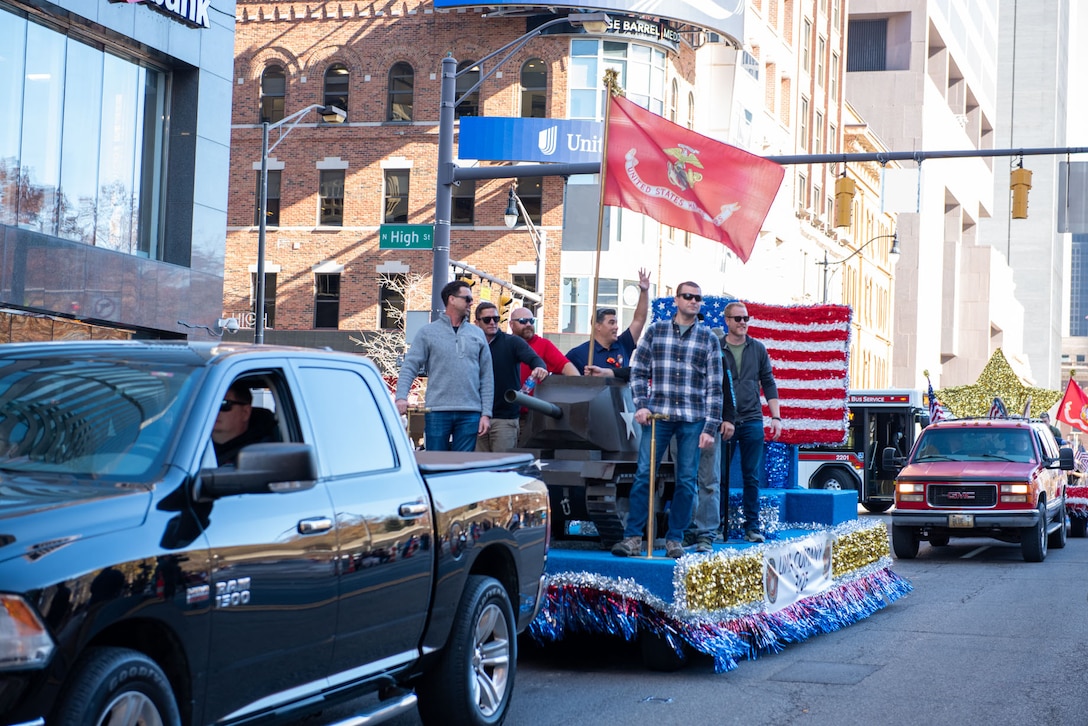 A dark pickup truck pulls a decorated float with several people on it. It has a US Flag in the background and patriotic colors.