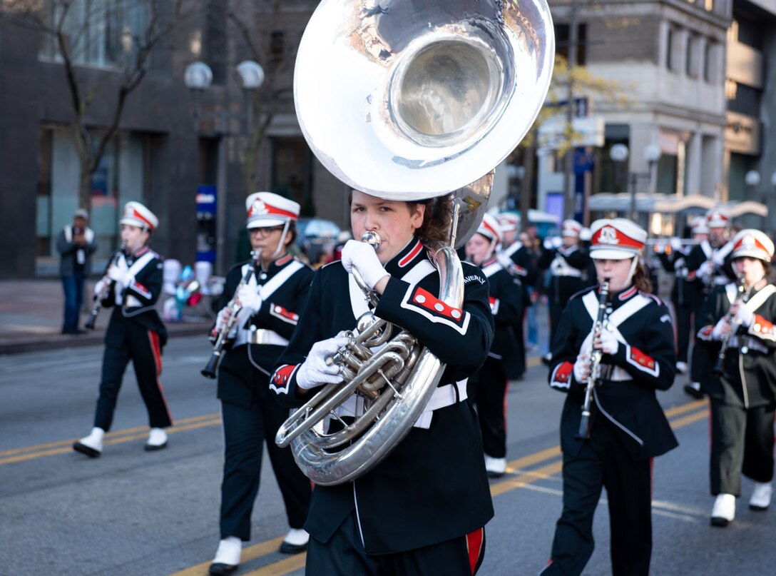 A member of a marching band in red, white and black march down a street in between high-rise buildings.