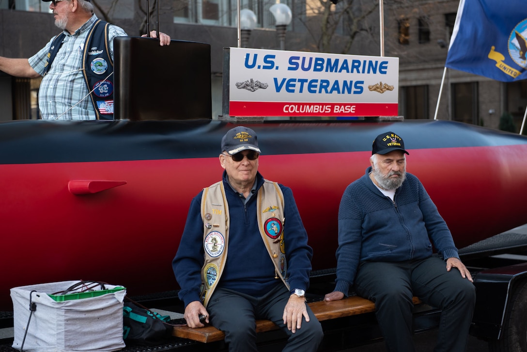 Two people in wearing military baseball caps sit on the side of a float.