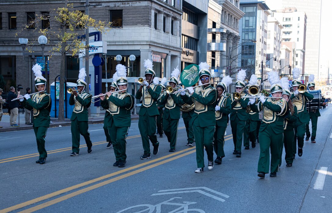 A marching band in green and gold march down a street in between high-rise buildings.