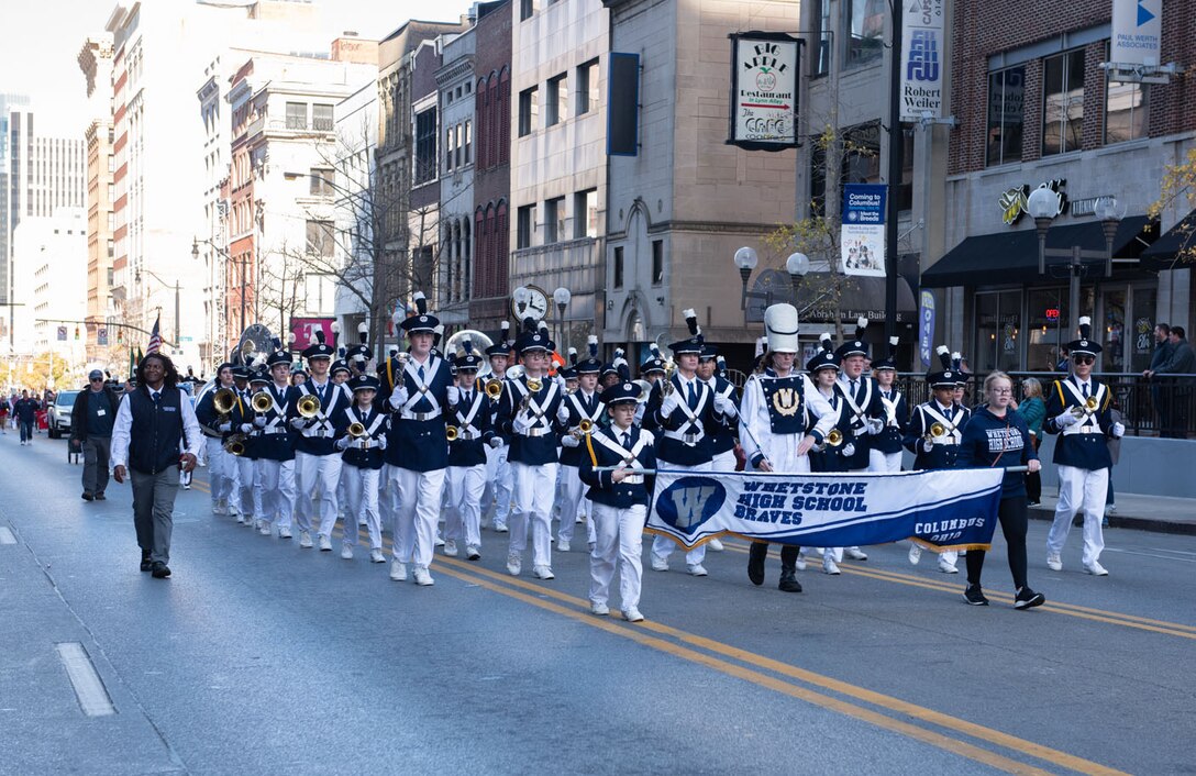 A marching band in blue and white march down a street in between high-rise buildings.