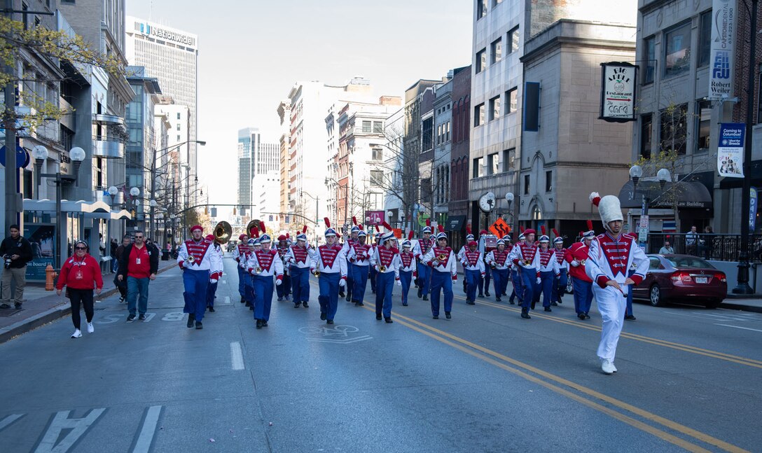 A marching band in red, white and blue march down a street in between high-rise buildings.