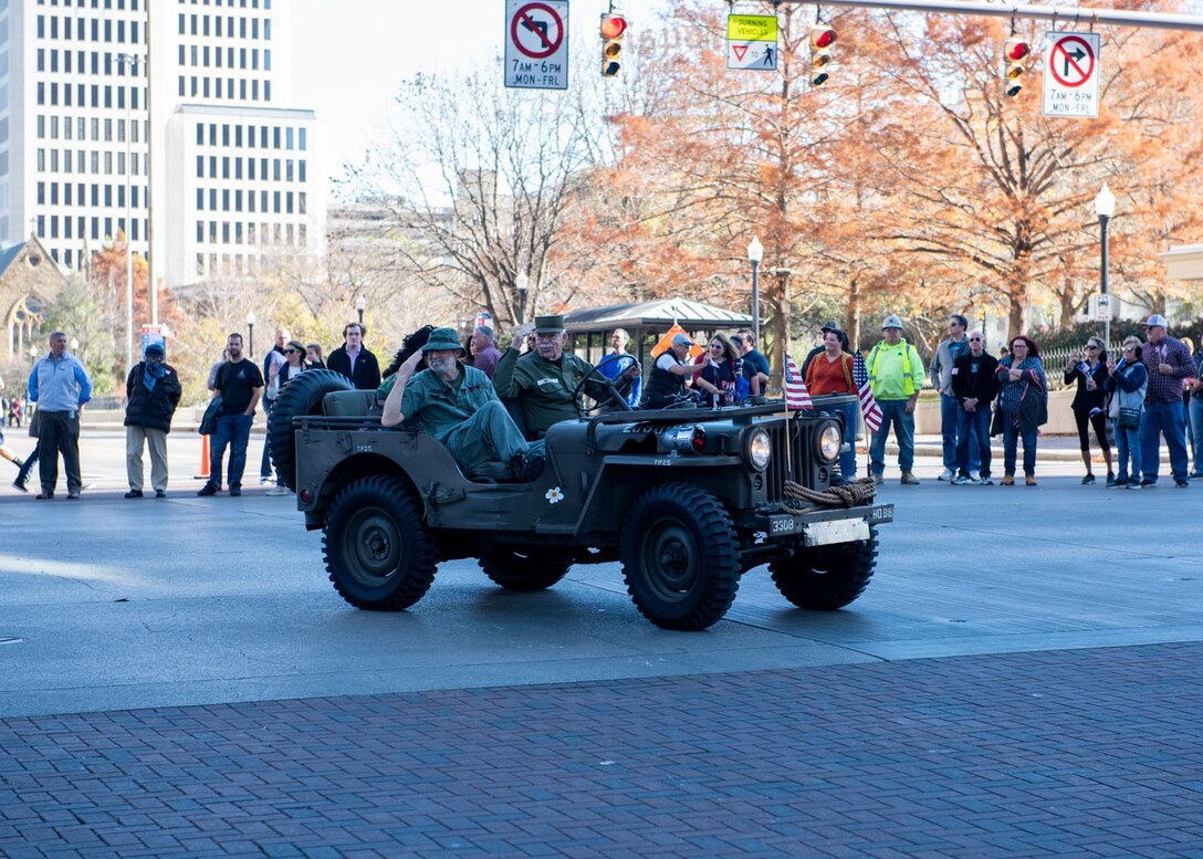 Two men in a WWII era Jeep turn a corner and salute on a street in between high rise buildings.