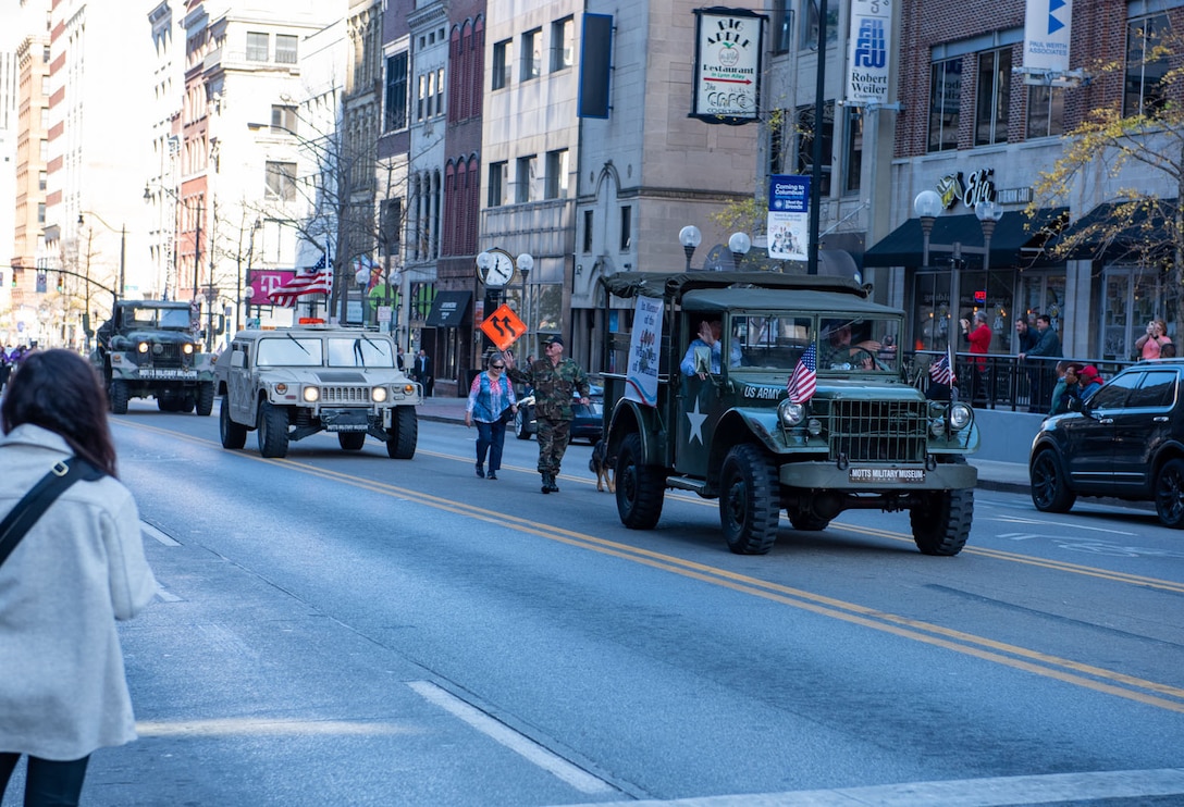 Military vehicles drive down a road in between high-rise buildings.