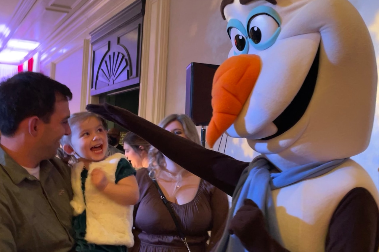 A life-sized snowman pats the head of a little girl in a man’s arms.