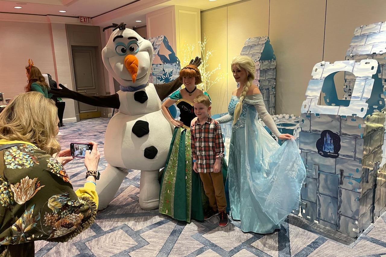 A boy stands beside two women dressed as princesses and a life-sized snowman.