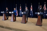Three men and one woman stand abreast of one another behind lecterns.