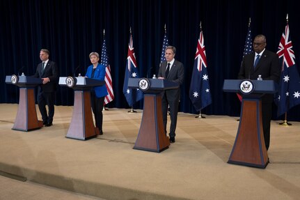 Three men and one woman stand behind lecterns with flags in the background.