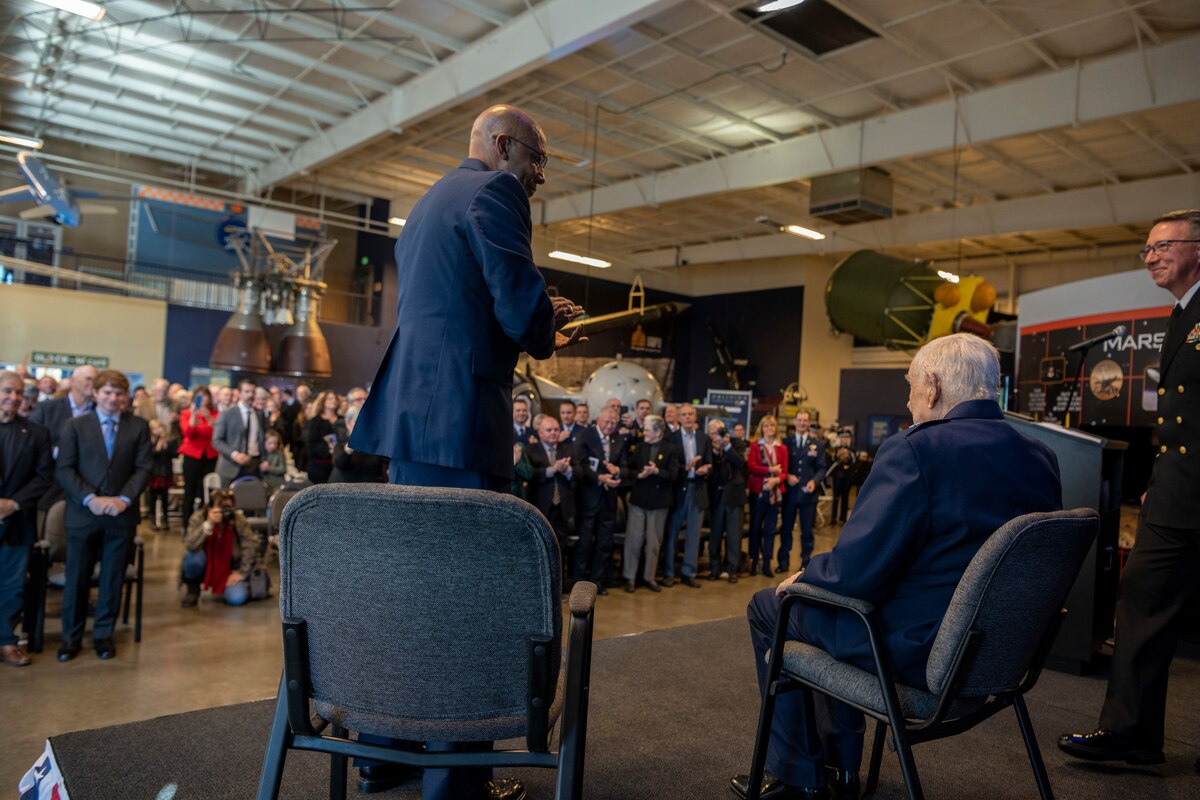 And older man sits on the stage as everyone around him stands and claps in celebration of him.