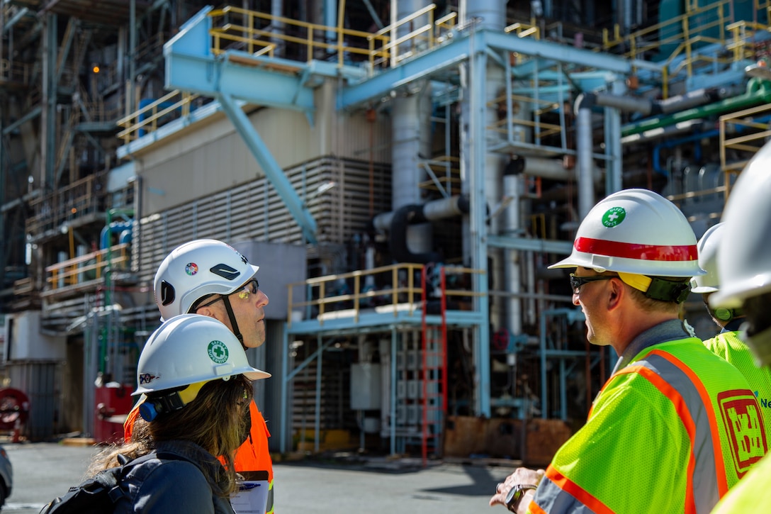 Three individuals look at the exterior of a power generation facility. The individuals are wearing safety gear.