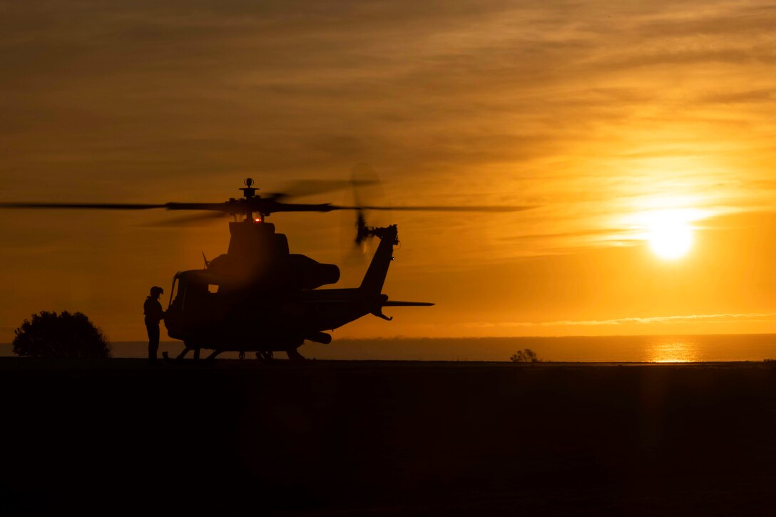 A Marine stands next to a helicopter near a body of water under a sunlit sky.