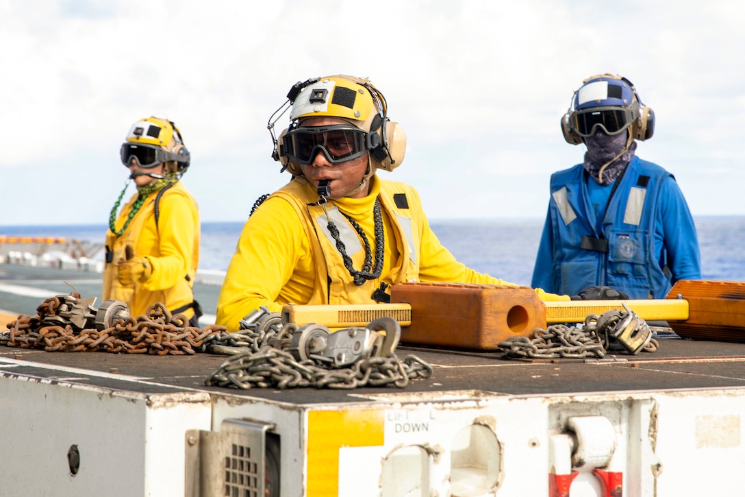 Three sailors stand next to a two truck aboard a ship at sea.