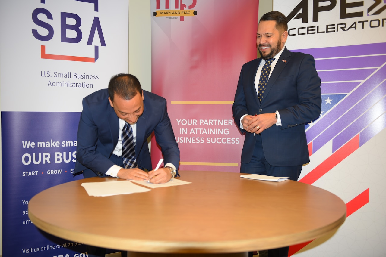 A man leans over a table and signs a document as another man watches.