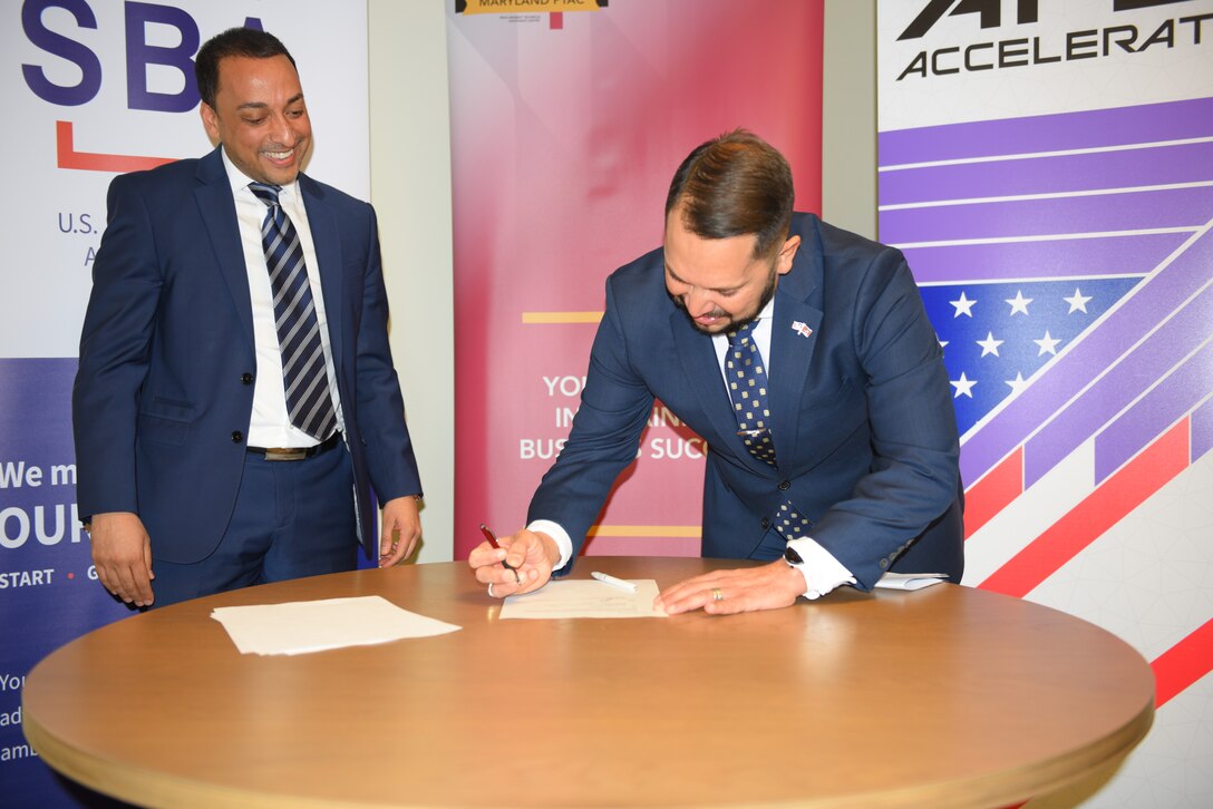 A man leans over a table and signs a document as another man watches.