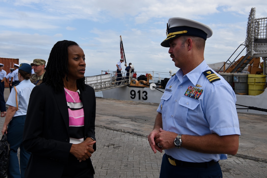 A man and woman talk while standing outside; a ship is in the background.