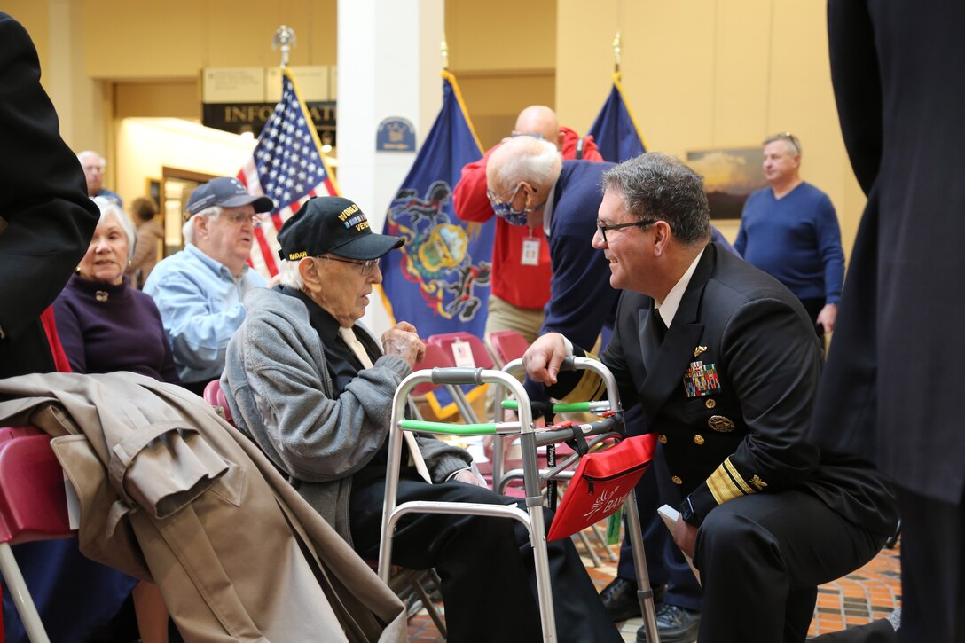 A smiling sailor kneels and talks to a seated veteran in a flag-adorned room.