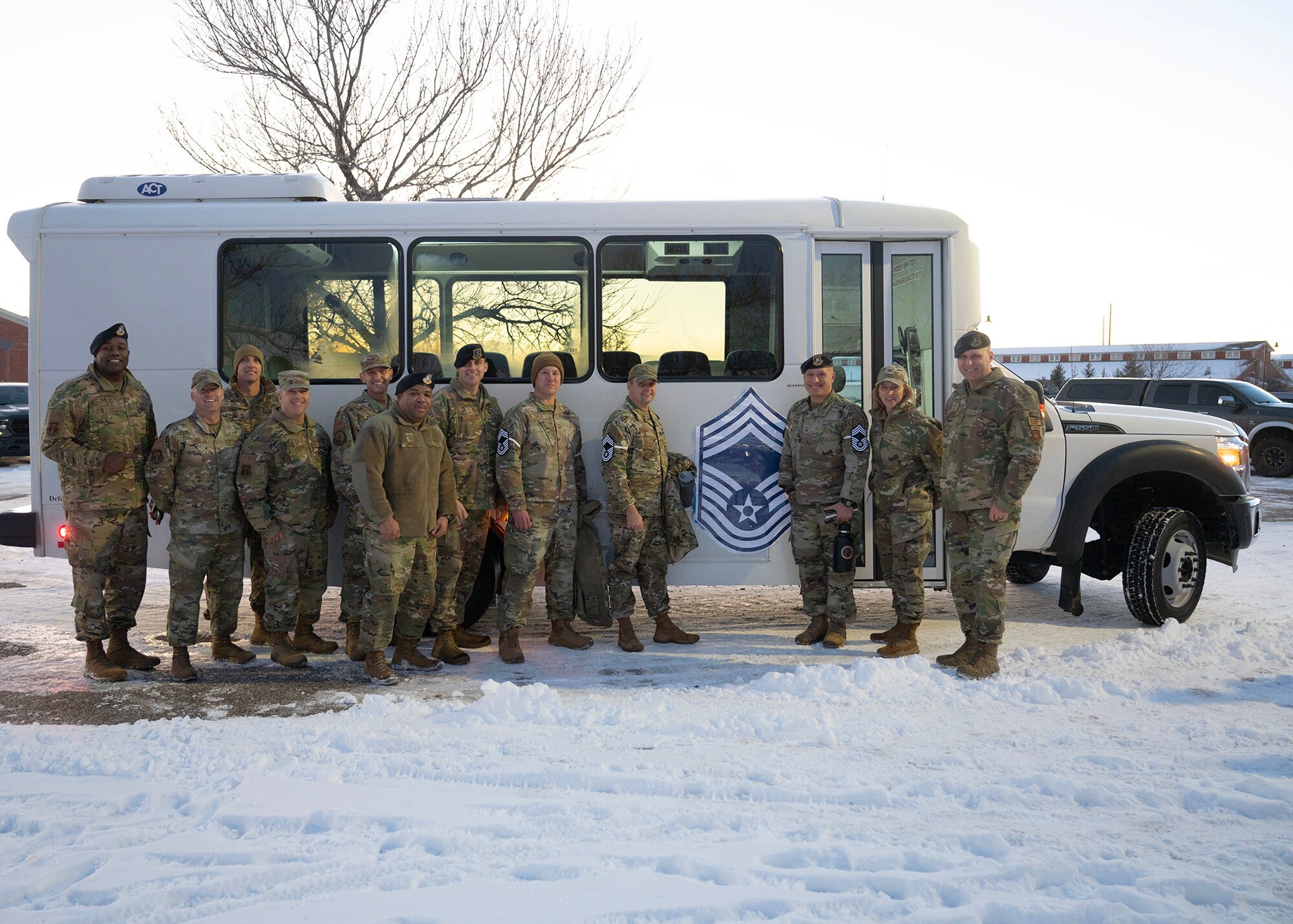 12 members of the Mighty Ninety standing in the snow in front of a bus adorned with a Chief Master Sergeant rank badge