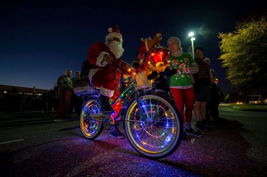 A soldier dressed as Santa sits on a bike with colored lights in the spokes of its wheels at night.