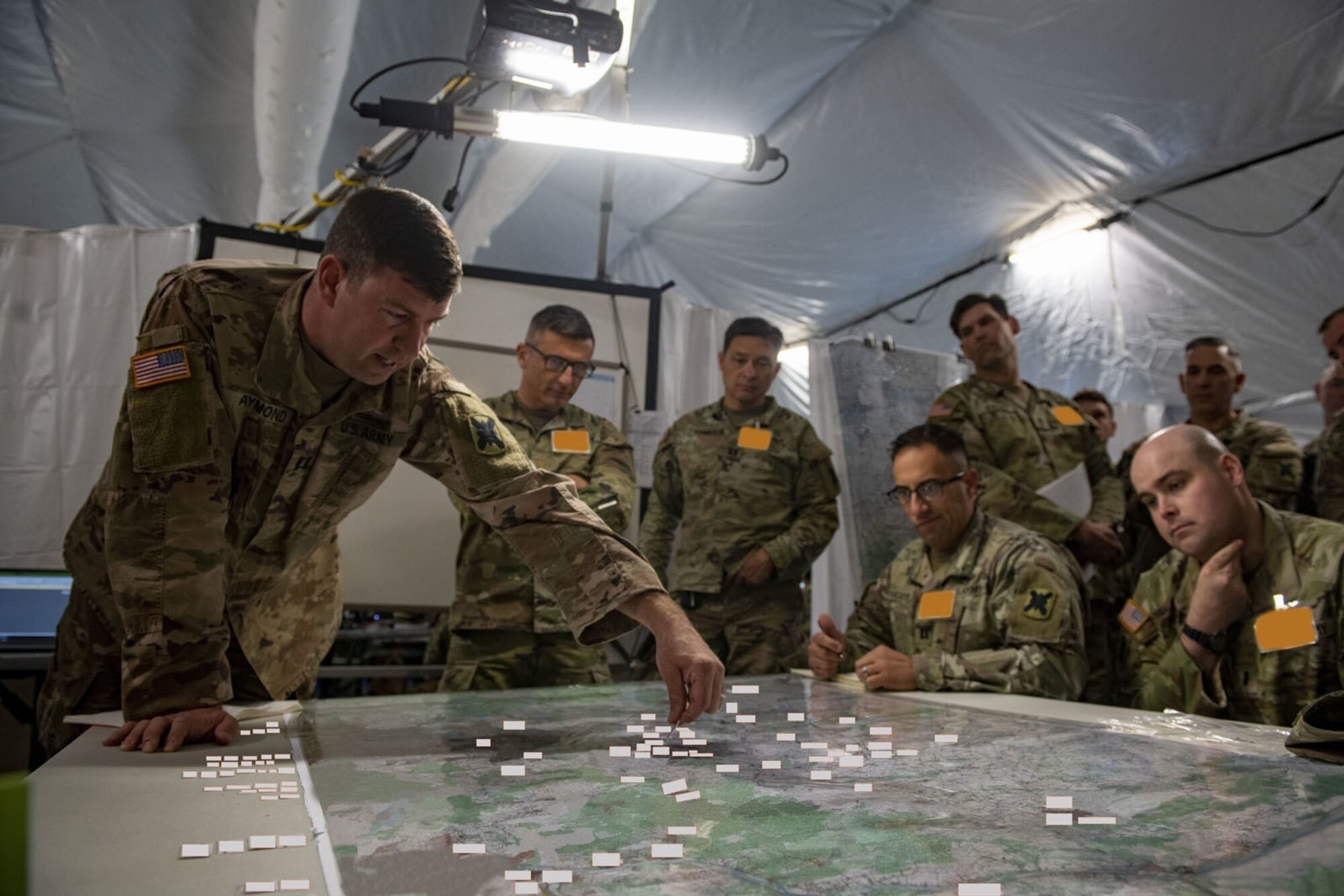 seven military members sitting and standing reviewing unit movements on table chart in a tent
