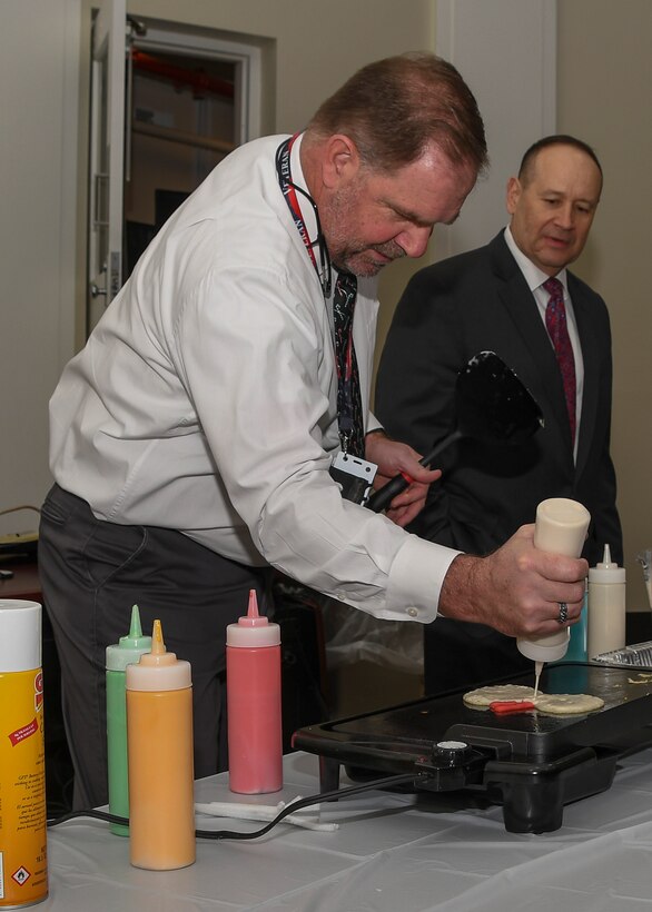 Man making pancakes using squeeze bottle as another man observes.