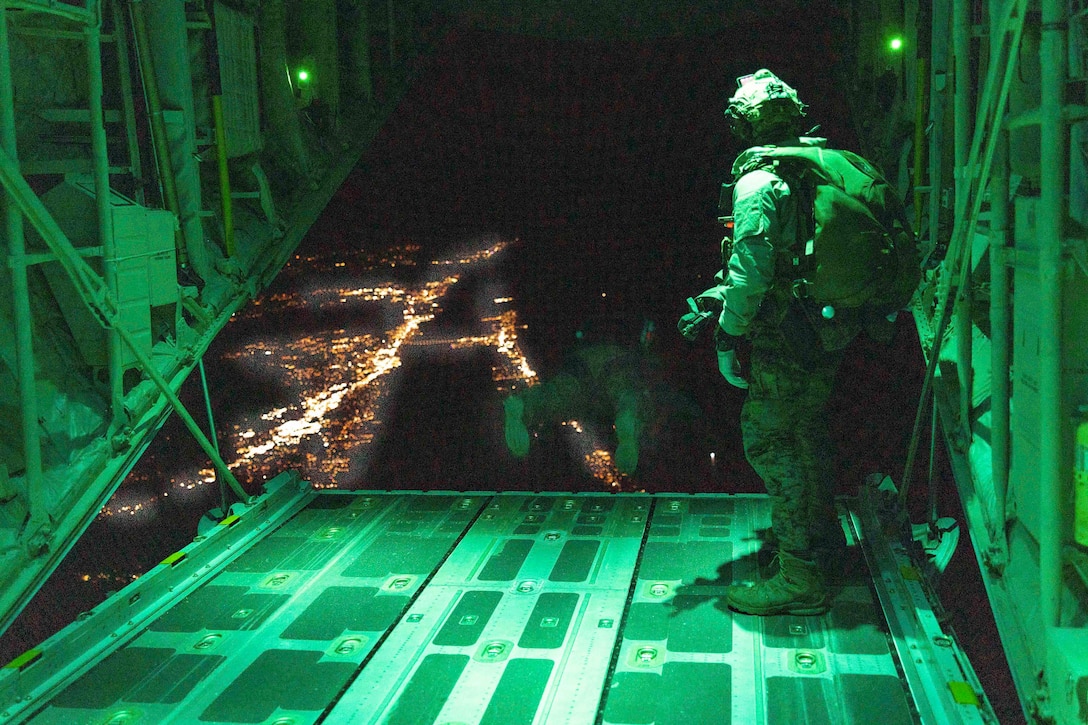 A Marine jumps out of an aircraft over a city as a fellow Marine watches.