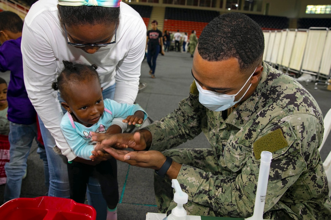 A sailor wearing a face mask washes a child’s hands at a medical site.