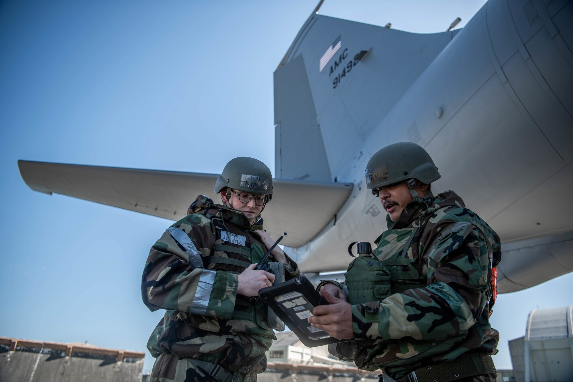 Two Airmen wearing mission oriented protective posture gear look at a checklist under the tail of an aircraft.