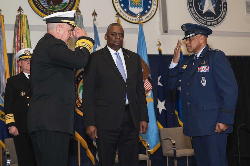 A Navy admiral and Air Force general salute each other on stage as Lloyd J. Austin III stands between them.