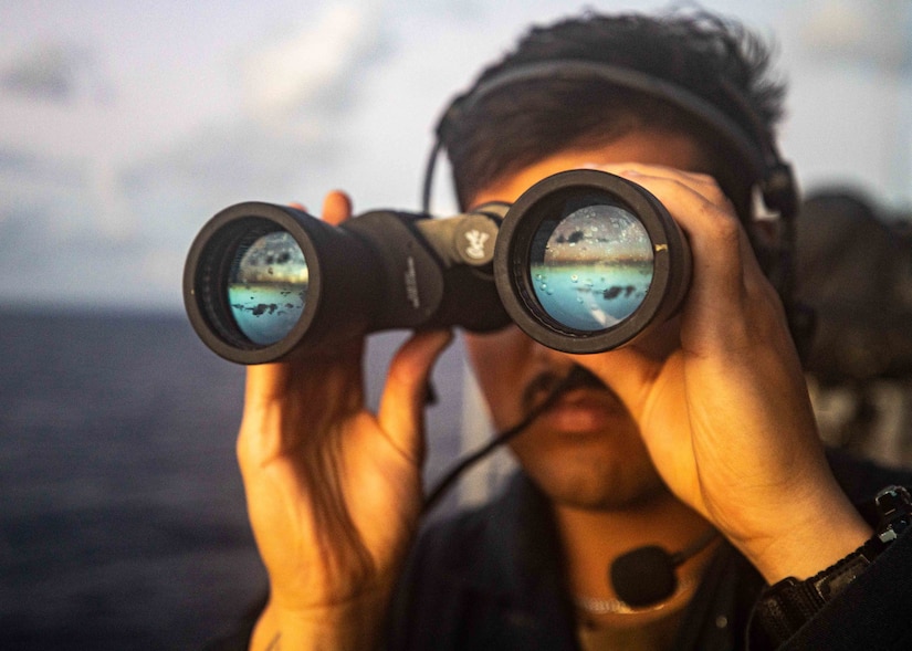 A sailor holds binoculars to his face.