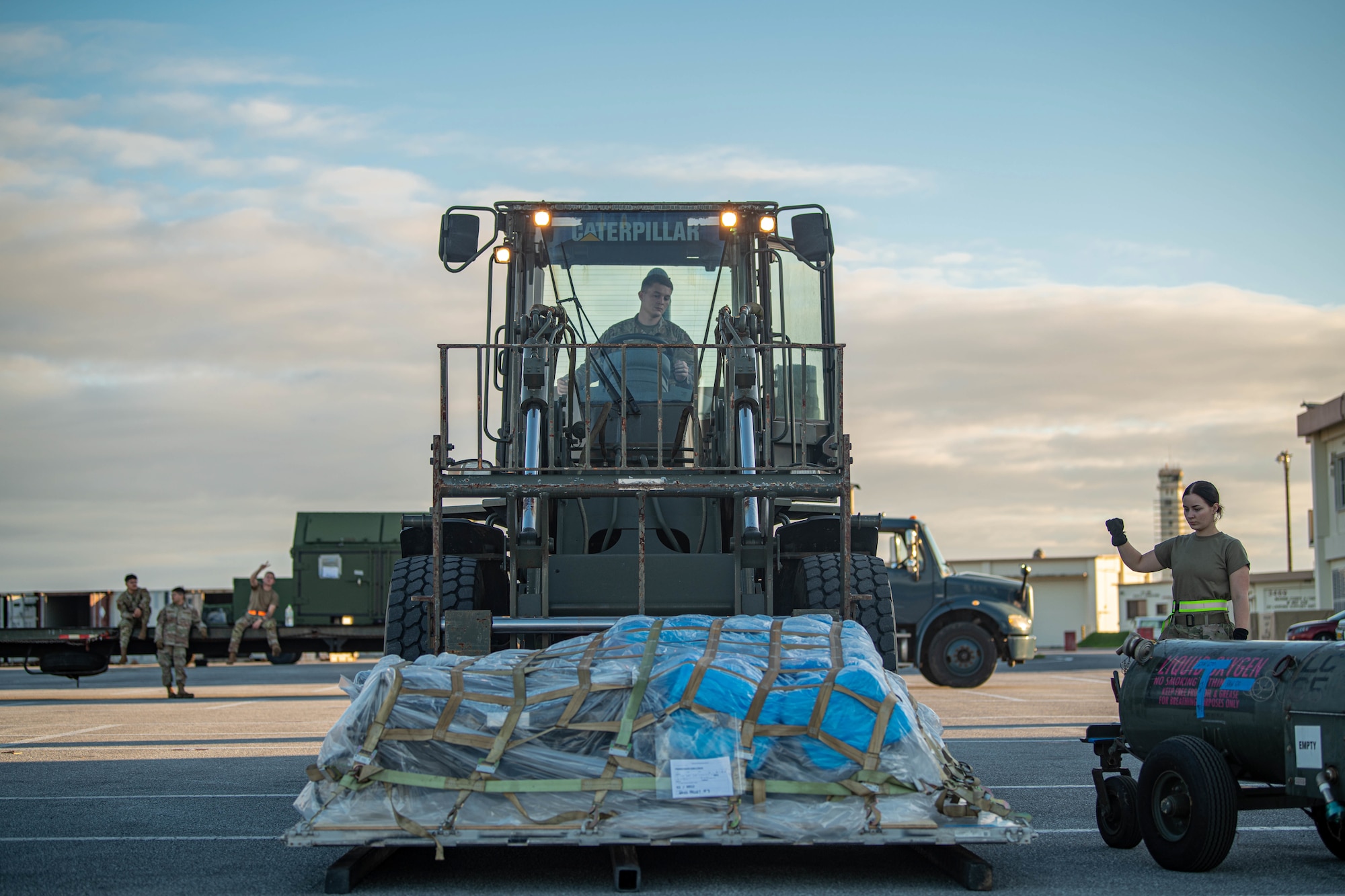 One Airman drives a fork lift with a pallet on it while another Airman guides them.