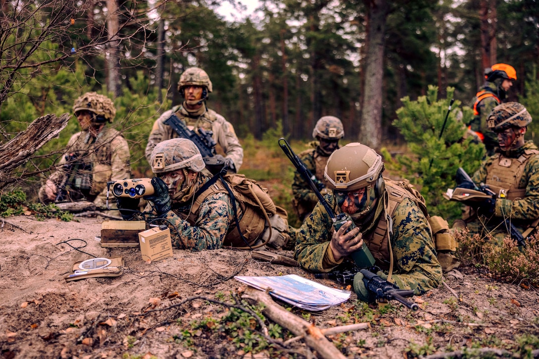 A Marine lying on ground looks through binoculars while  another speaks into a handheld device and others watch.