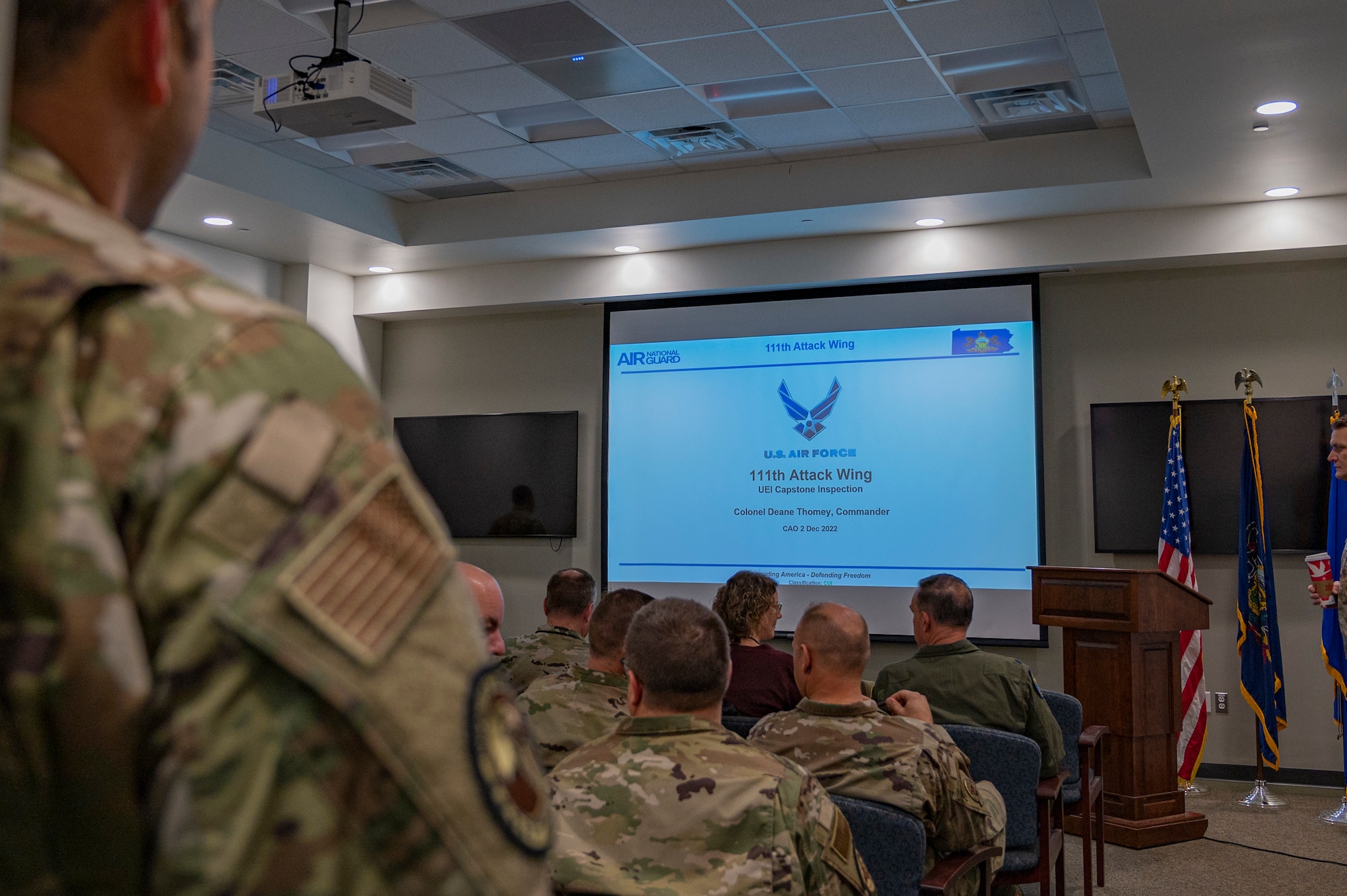 View over the shoulder of a uniformed military member of a group of assembled military and civilian personnel.