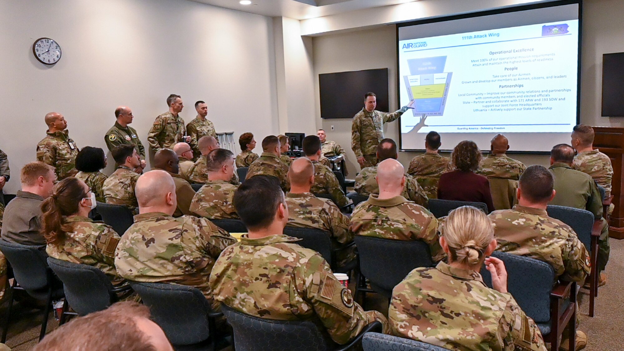 Uniformed military man gestures toward briefing slide while addressing assembled group of military and civilian personnel.