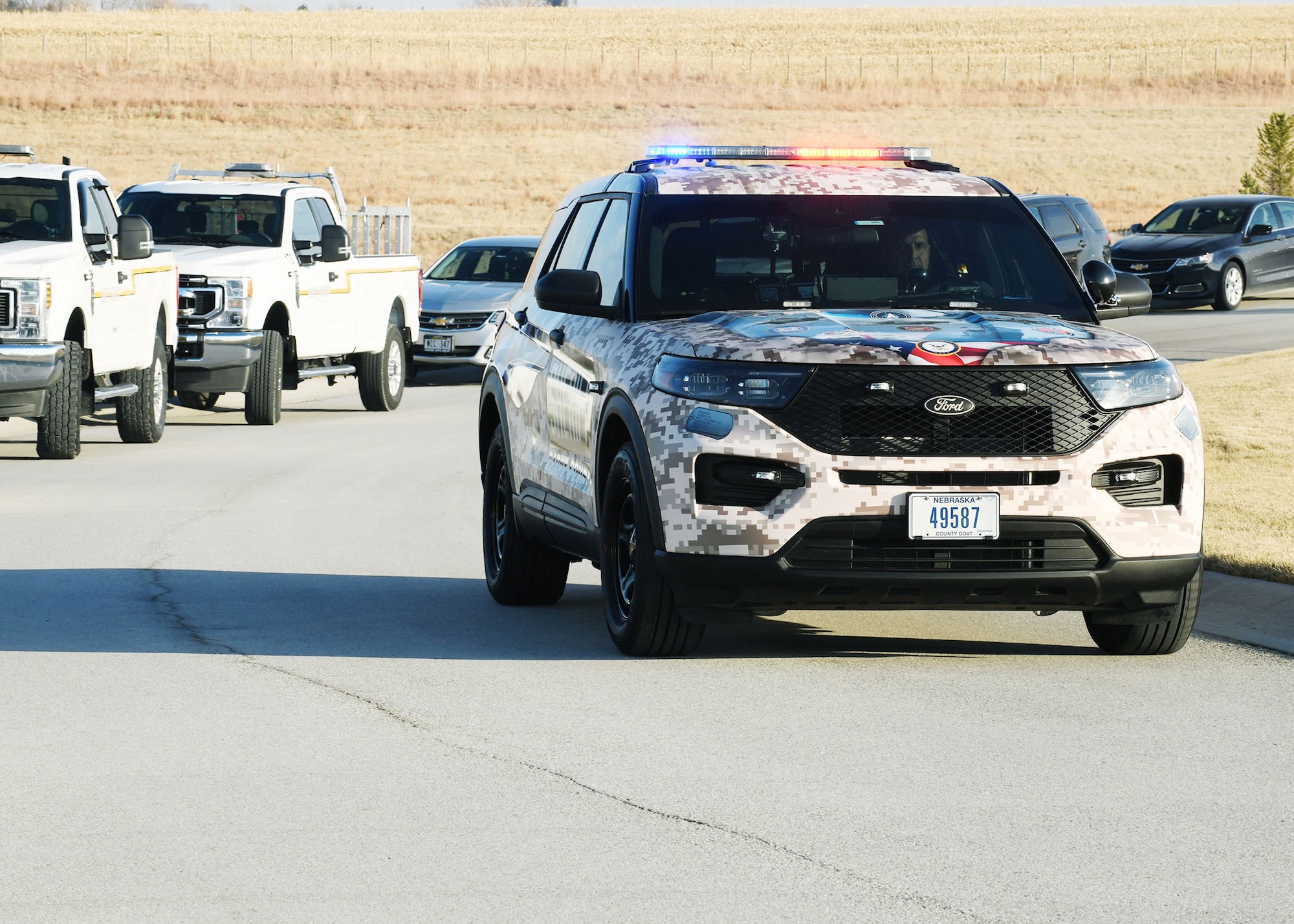 law enforcement car with flashing lights covered in military style paint and dog tags drives through cemetery.