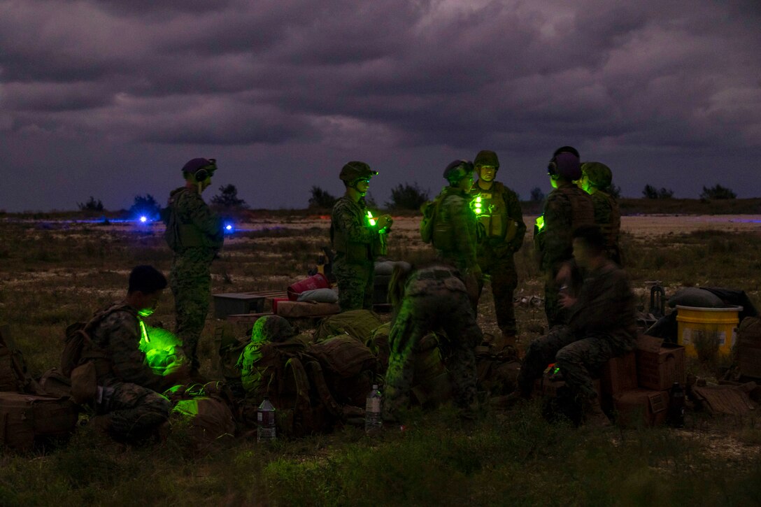 Marines gather in a field in the dark illuminated by yellow and blue lights.