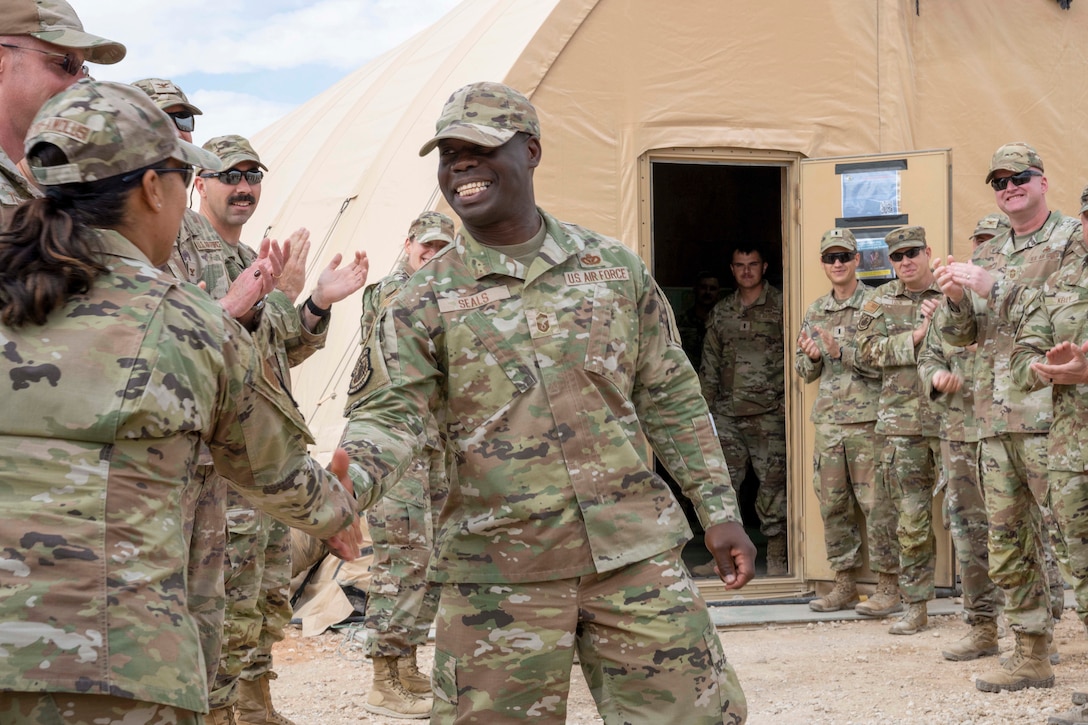 An airman shakes hands with a fellow airman as other airmen applaud in front of a tent.